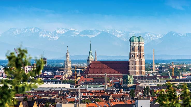 München im Sommer mit Frauenkirche und Berge im Hintergrund.